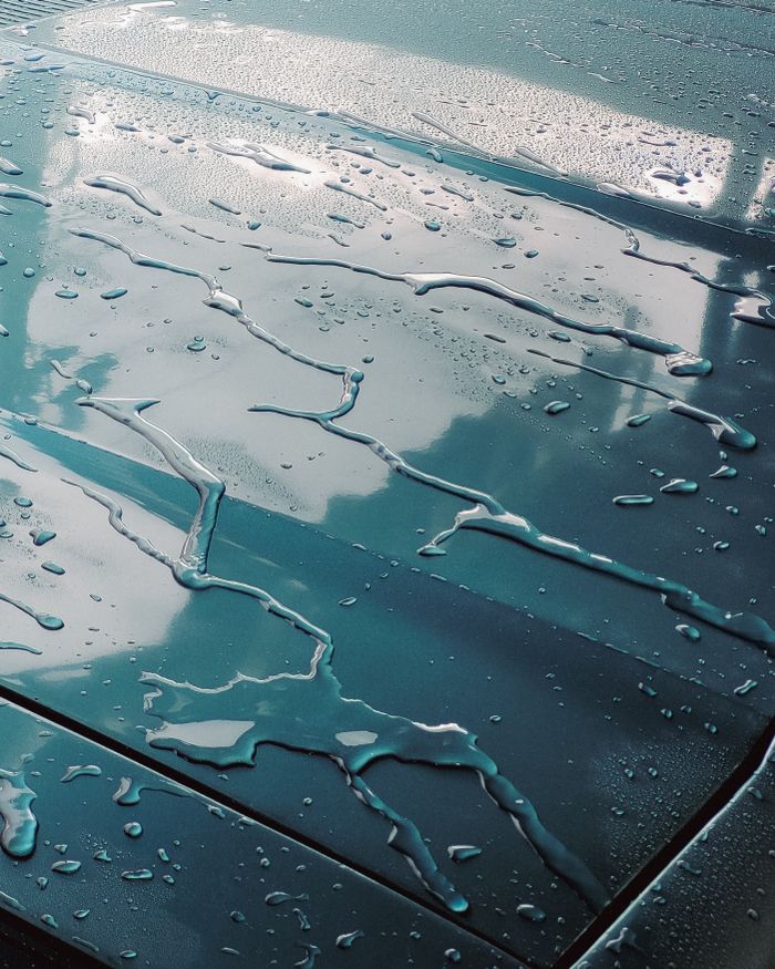 Close-up of water droplets on car hood reflecting clouds and sky, creating a serene pattern.