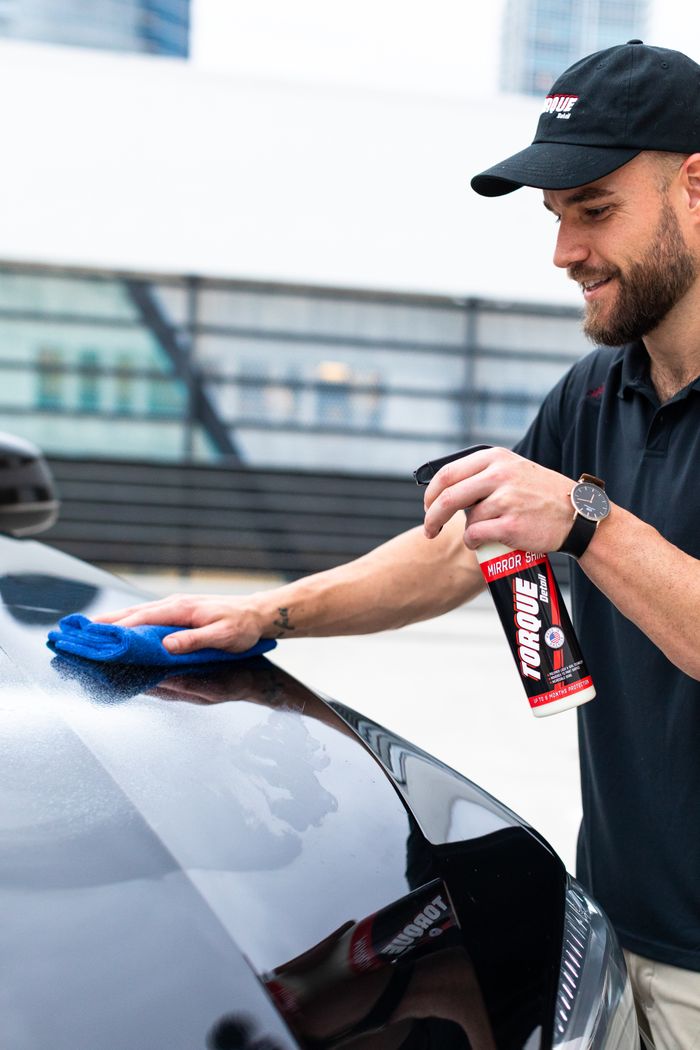 Man wiping down a car exterior with cleaning spray on a sunny day.